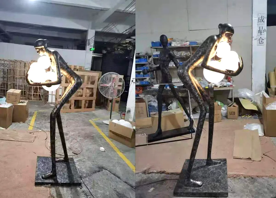 Bronze figure sculpture floor lamps holding glowing glass orbs — a pair on the factory floor